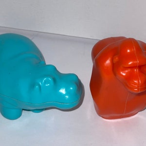 May include: Two plastic toy animals, a blue hippopotamus and an orange gorilla. The hippopotamus is lying on its belly with its legs tucked underneath it. The gorilla is sitting upright with its arms crossed in front of it.