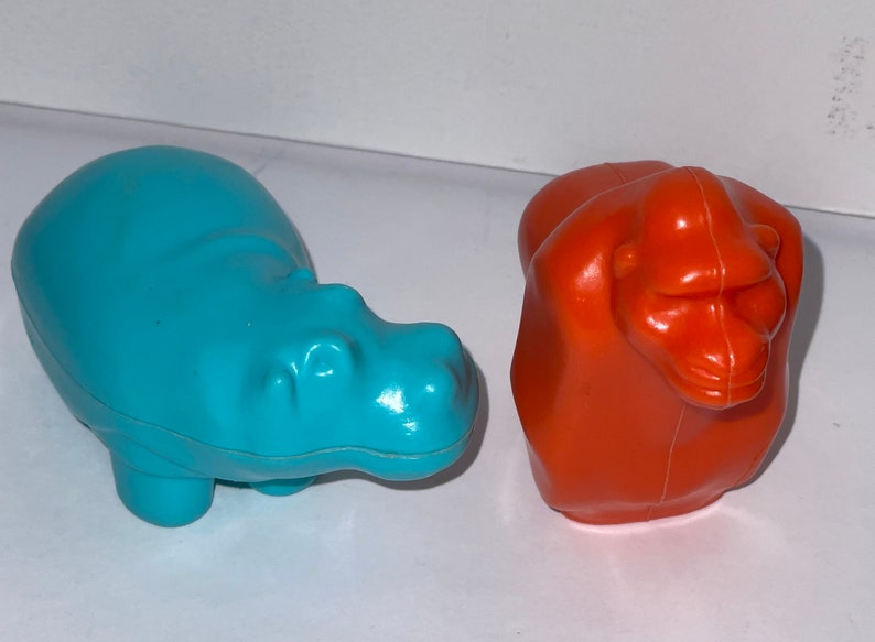 May include: Two plastic toy animals, a blue hippopotamus and an orange gorilla. The hippopotamus is lying on its belly with its legs tucked underneath it. The gorilla is sitting upright with its arms crossed in front of it.