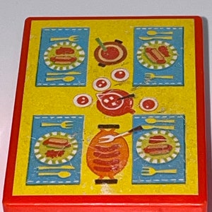 Pu&ograve; includere: Un tavolo da gioco in plastica gialla con bordo rosso. Il tavolo ha una tovaglia a quadri blu e bianchi con un disegno di hot dog, piatti, forchette, cucchiai e una pentola di cibo.