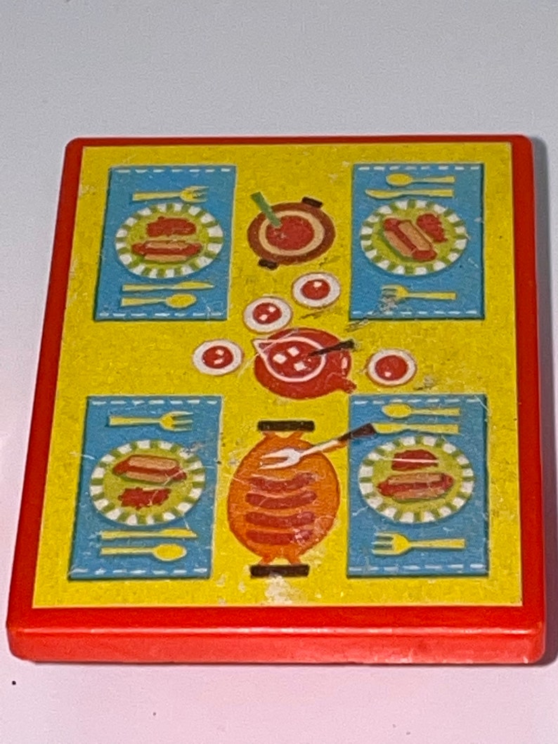 Pu&ograve; includere: Un tavolo da gioco in plastica gialla con bordo rosso. Il tavolo ha una tovaglia a quadri blu e bianchi con un disegno di hot dog, piatti, forchette, cucchiai e una pentola di cibo.