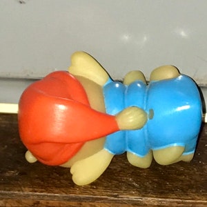 Puede incluir: Una peque&ntilde;a figura de pl&aacute;stico azul claro y naranja de un personaje durmiendo con un gorro rojo. La figura est&aacute; tumbada de lado con la cabeza apoyada en el brazo.