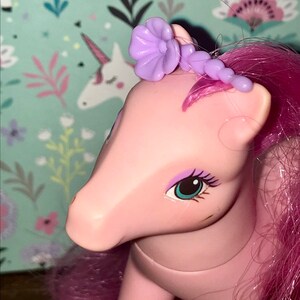 Puede incluir: Juguete de pony rosa con pelo morado y un accesorio para el pelo con forma de flor morada. El pony tiene ojos azules y una nariz rosa.