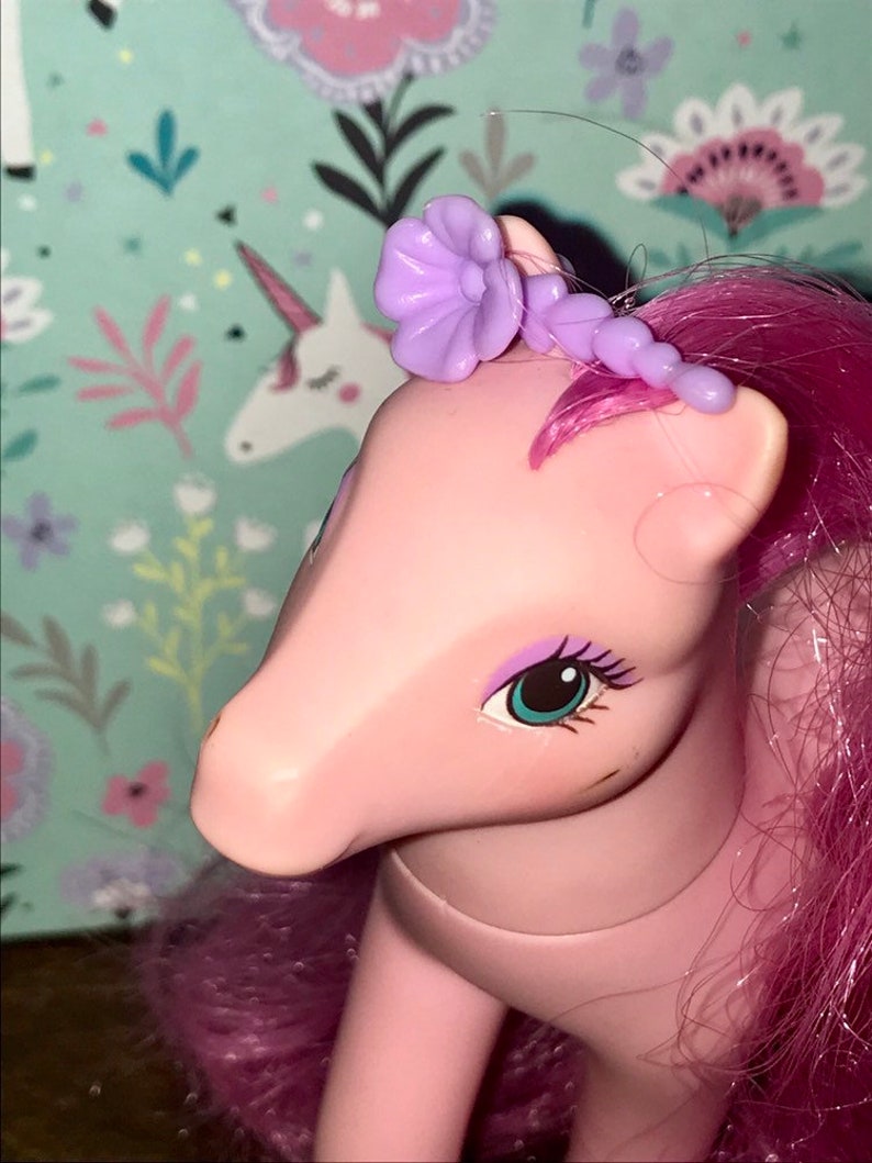 Puede incluir: Juguete de pony rosa con pelo morado y un accesorio para el pelo con forma de flor morada. El pony tiene ojos azules y una nariz rosa.