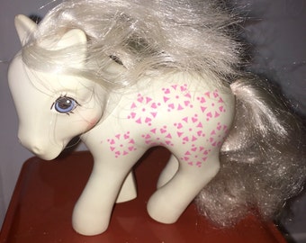 Może przedstawiać: Biały kucyk My Little Pony z wzorem różowych i białych serc na ciele. Kucyk ma długą, falującą białą grzywę i ogon.
