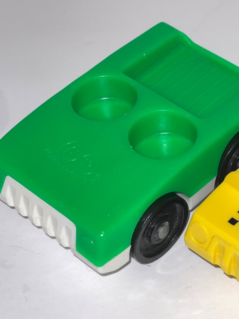 May include: Green toy truck with a white bumper and black wheels. The truck has two round indentations on the bed and a rectangular indentation with a ribbed surface. The truck is made of plastic.