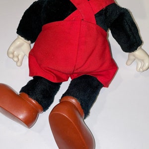 May include: A black plush Mickey Mouse toy wearing red overalls and brown shoes. The toy is lying on its back with its arms outstretched.