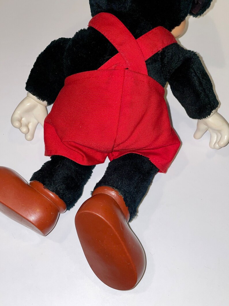 May include: A black plush Mickey Mouse toy wearing red overalls and brown shoes. The toy is lying on its back with its arms outstretched.