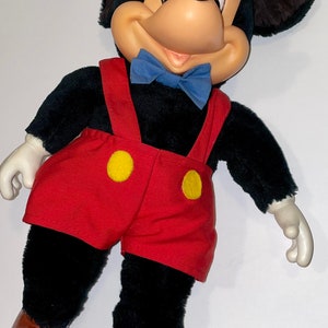 May include: A plush Mickey Mouse toy with black fur, red shorts with yellow buttons, and a blue bow tie. The toy has brown shoes and large, black ears.