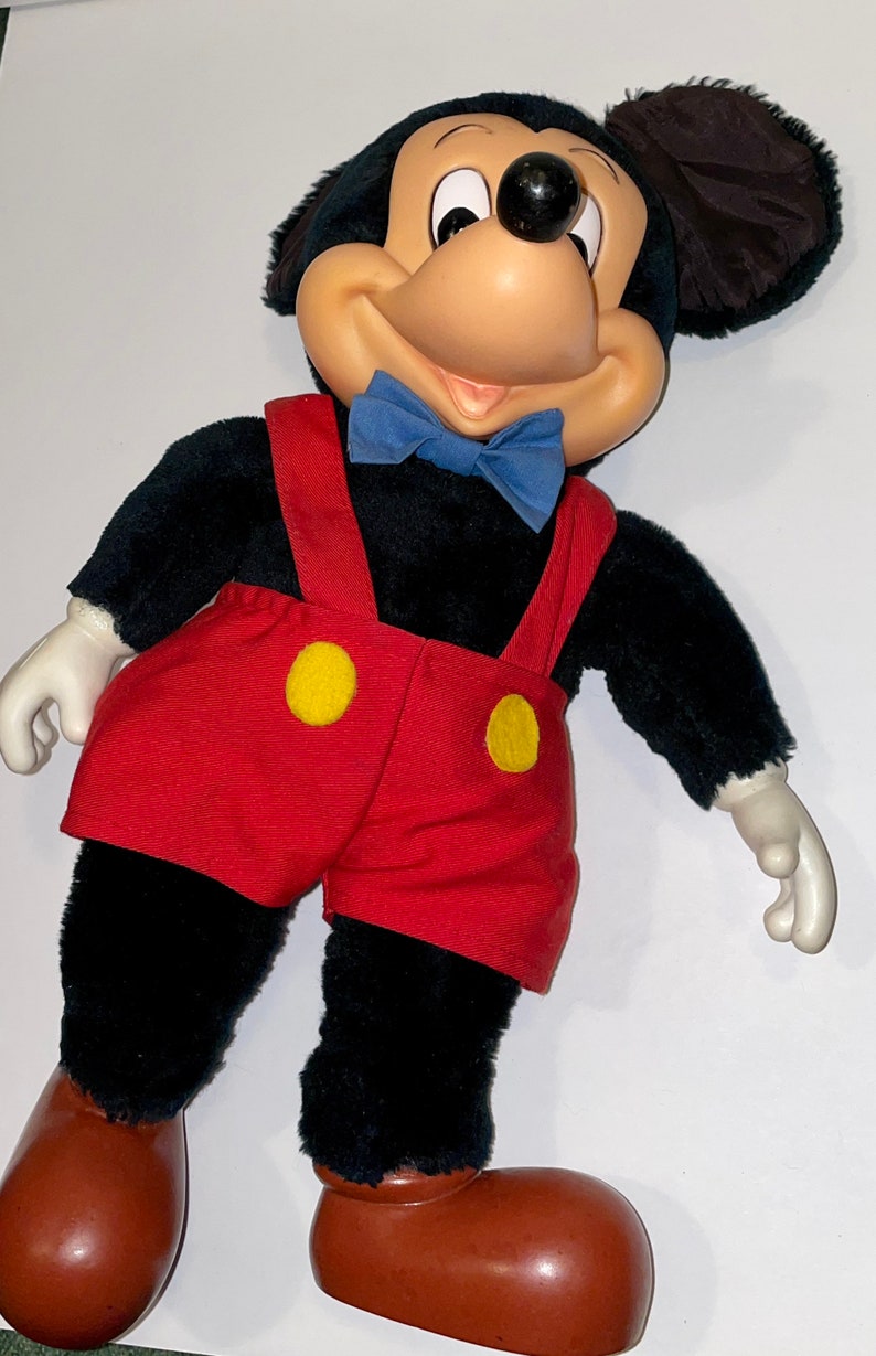 May include: A plush Mickey Mouse toy with black fur, red shorts with yellow buttons, and a blue bow tie. The toy has brown shoes and large, black ears.