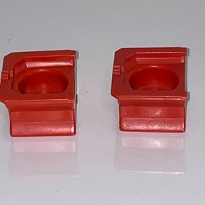 May include: Two small, red plastic containers with a square base and a round, indented top. The containers are made of a smooth, glossy plastic.