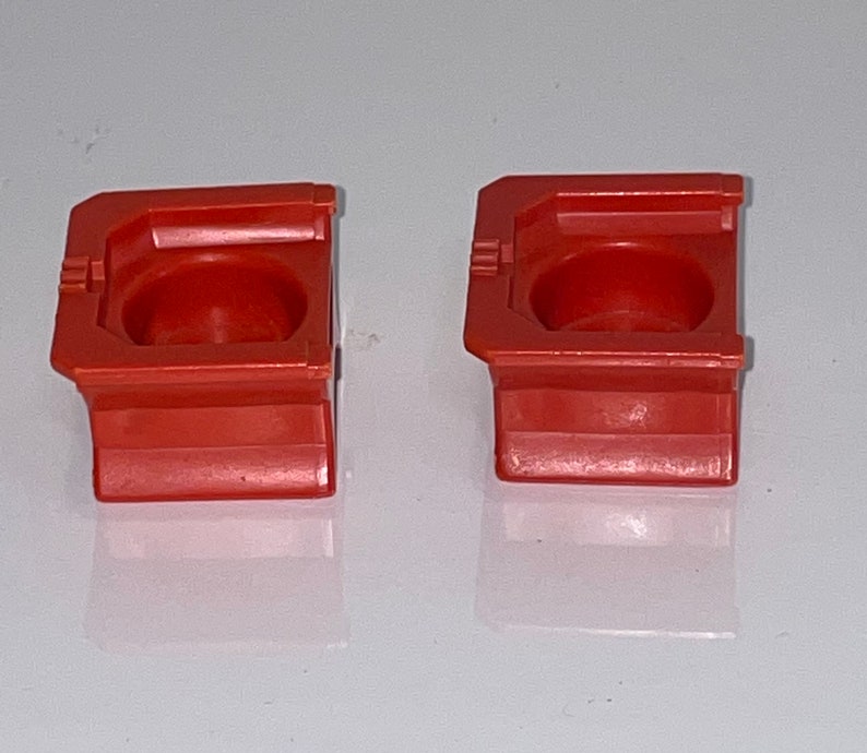 May include: Two small, red plastic containers with a square base and a round, indented top. The containers are made of a smooth, glossy plastic.
