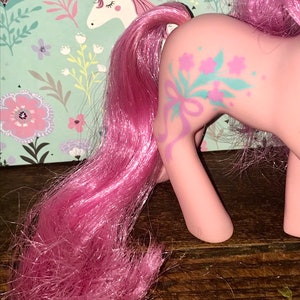 Puede incluir: Juguete de pony rosa con una larga melena y cola rosas. El pony tiene un dise&ntilde;o floral en un lado con una cinta azul.