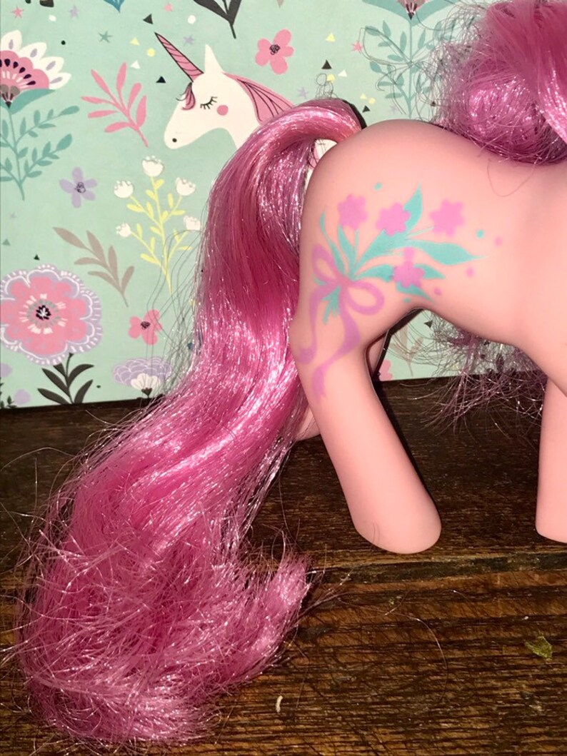 Puede incluir: Juguete de pony rosa con una larga melena y cola rosas. El pony tiene un dise&ntilde;o floral en un lado con una cinta azul.