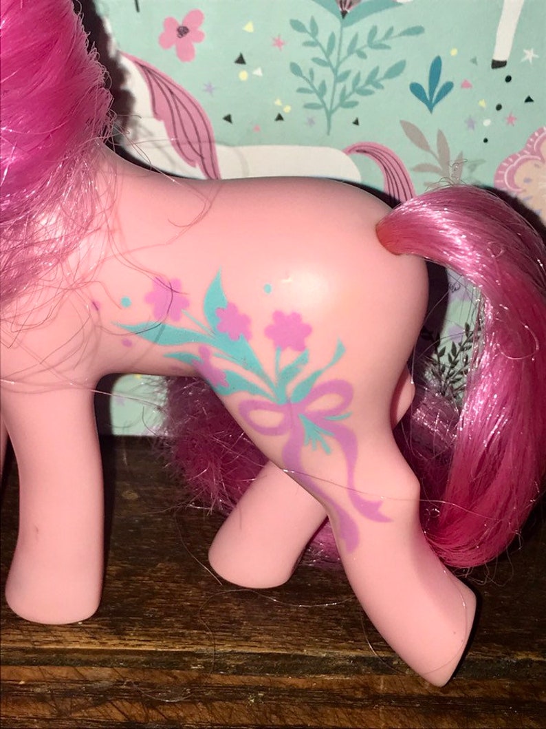 Puede incluir: Pony rosa My Little Pony con un dise&ntilde;o floral morado y azul en un lado. El pony tiene una melena y cola rosadas.