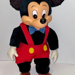 May include: A plush Mickey Mouse toy with black fur, red shorts with yellow buttons, and brown shoes. The toy has a blue bow tie and a friendly expression.