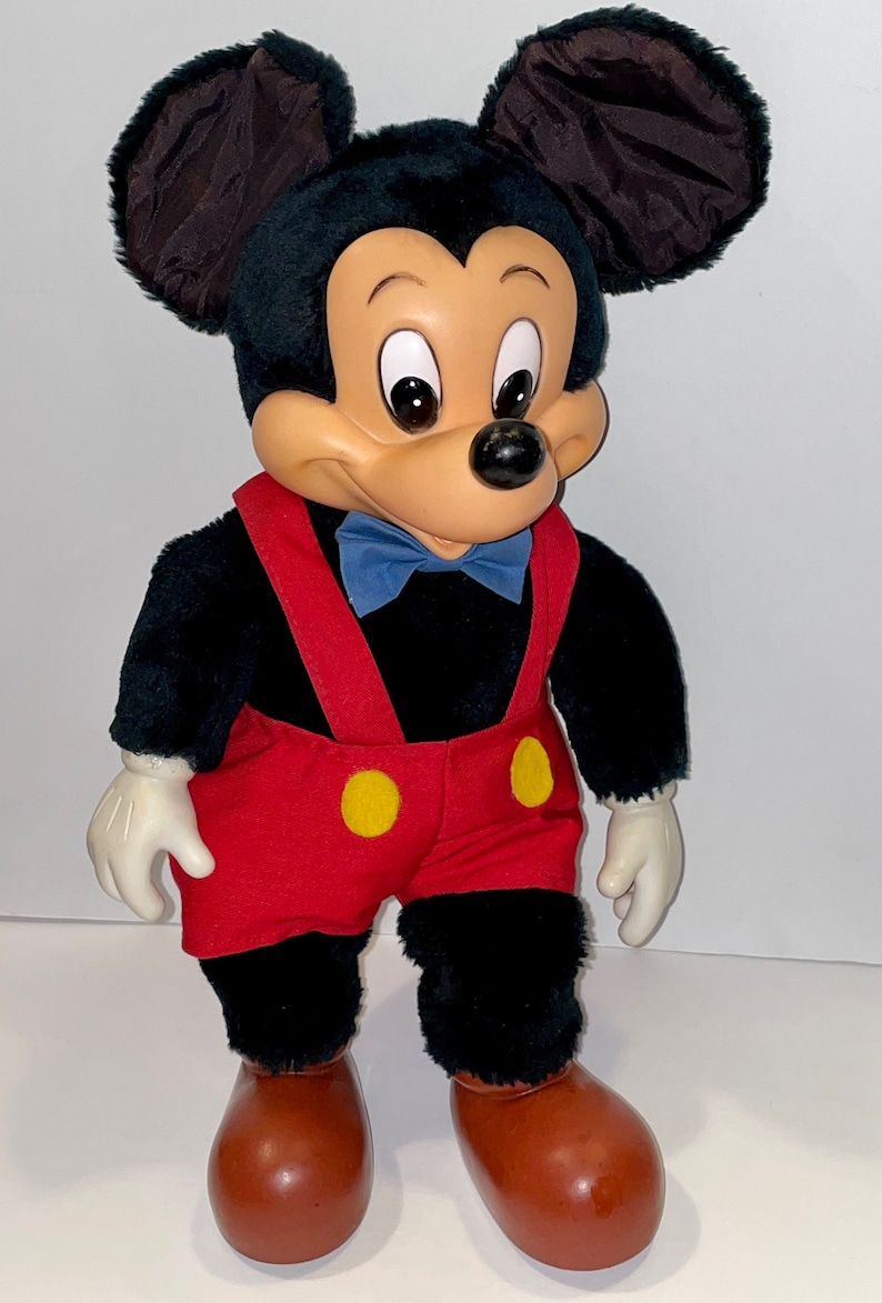 May include: A plush Mickey Mouse toy with black fur, red shorts with yellow buttons, and brown shoes. The toy has a blue bow tie and a friendly expression.