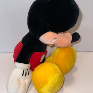 May include: A plush Mickey Mouse toy sitting with its head down. The toy is black with red shorts and yellow shoes.