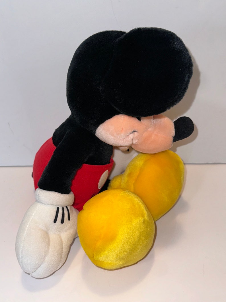 May include: A plush Mickey Mouse toy sitting with its head down. The toy is black with red shorts and yellow shoes.
