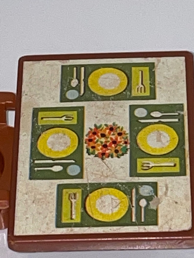 May include: A brown plastic play table with a white and brown marbled surface. The table has a green and yellow design with four place settings, each with a plate, fork, knife, and spoon. A floral centerpiece is in the center of the table.