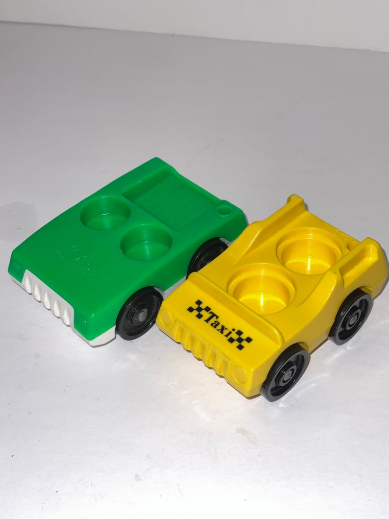 May include: Two plastic toy cars, one green and one yellow. The yellow car has "Taxi" printed on the side. Both cars have two cup holders.