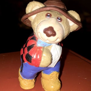 May include: A brown teddy bear wearing a brown hat, a red and black plaid shirt, blue jeans, and brown boots. The bear is standing on a red surface.