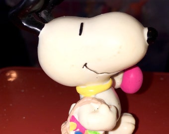 May include: A white plastic figurine of Snoopy holding a pink basket filled with colorful Easter eggs. Snoopy is wearing a yellow collar and has black ears.