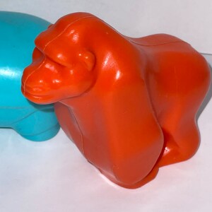 May include: Orange plastic toy gorilla. The gorilla is standing on a white surface.