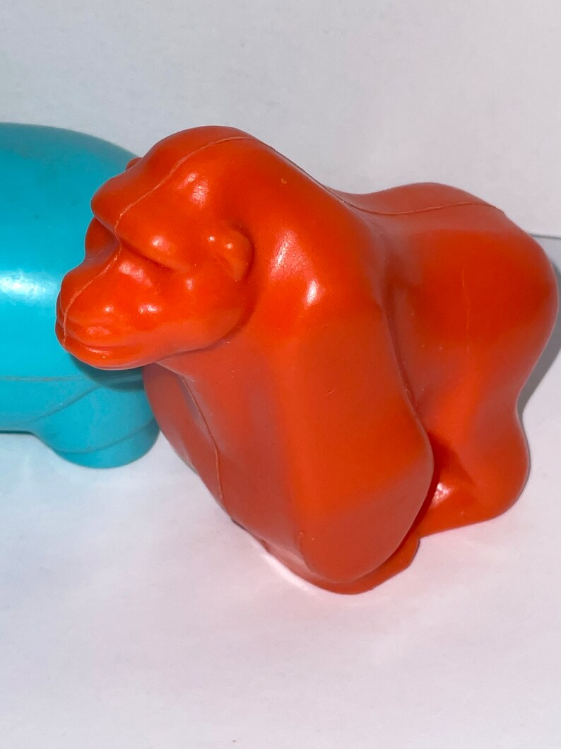 May include: Orange plastic toy gorilla. The gorilla is standing on a white surface.