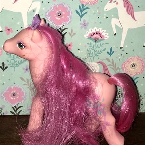 Puede incluir: Pony rosa My Little Pony con un dise&ntilde;o floral morado y azul en su costado. El pony tiene una larga melena y cola rosadas. Tiene una corona morada con una gema azul en la parte superior.