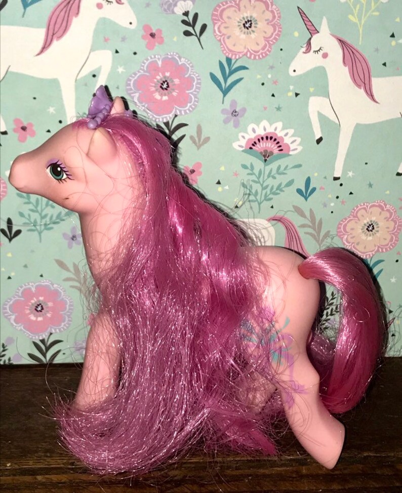 Puede incluir: Pony rosa My Little Pony con un dise&ntilde;o floral morado y azul en su costado. El pony tiene una larga melena y cola rosadas. Tiene una corona morada con una gema azul en la parte superior.