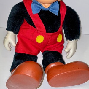 May include: A plush black Mickey Mouse toy wearing red overalls with yellow buttons, a blue bow tie, and brown shoes.