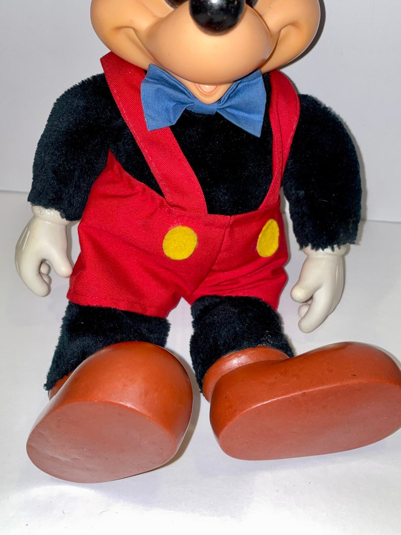 May include: A plush black Mickey Mouse toy wearing red overalls with yellow buttons, a blue bow tie, and brown shoes.