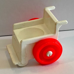 May include: White plastic toy wheelchair with a red wheel. The wheelchair has a seat and a back.
