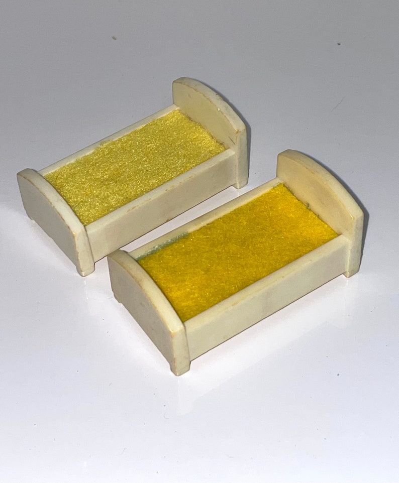 May include: Two white plastic doll beds with yellow fabric mattresses. The beds are small and would be suitable for a dollhouse.