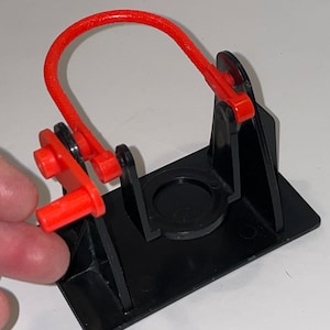 May include: A black plastic toy with a red loop and lever. The toy is designed to launch a small object.