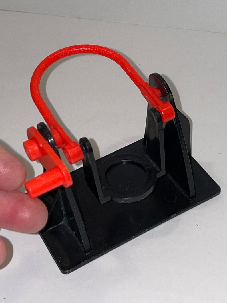 May include: A black plastic toy with a red loop and lever. The toy is designed to launch a small object.