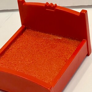 May include: A red ink pad with a textured surface. The ink pad has a rectangular shape with a curved back and a small decorative element on the top. The ink pad is designed for use with stamps.