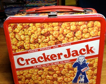 May include: A red metal lunchbox with a Cracker Jack logo and a cartoon sailor boy. The lunchbox has a white background with a yellow and brown pattern of popcorn and peanuts. The text on the lunchbox reads "CANDIED POPCORN AND PEANUTS Cracker Jack BRAND THE MORE YOU EAT THE MORE YOU WANT!"