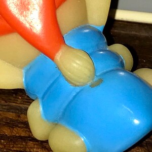 Puede incluir: Un juguete de goma vintage de color rojo, amarillo y azul. El juguete tiene forma de persona sentada con las piernas cruzadas. El juguete tiene un peque&ntilde;o agujero en el lateral.