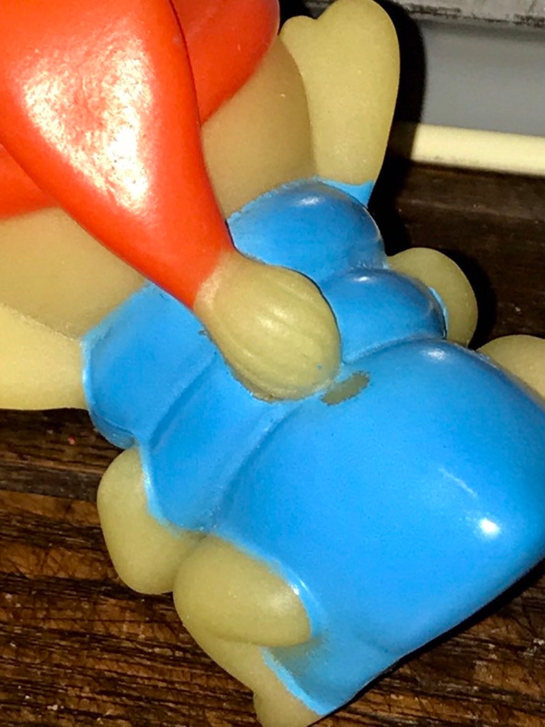 Puede incluir: Un juguete de goma vintage de color rojo, amarillo y azul. El juguete tiene forma de persona sentada con las piernas cruzadas. El juguete tiene un peque&ntilde;o agujero en el lateral.