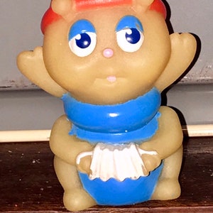 Puede incluir: Un juguete de goma amarillo con un sombrero rojo, un mono azul y un acorde&oacute;n blanco. El juguete tiene una cara amigable con ojos azules y una nariz rosa.