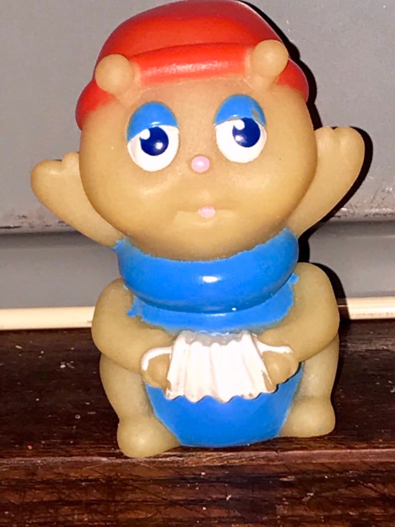 Puede incluir: Un juguete de goma amarillo con un sombrero rojo, un mono azul y un acorde&oacute;n blanco. El juguete tiene una cara amigable con ojos azules y una nariz rosa.