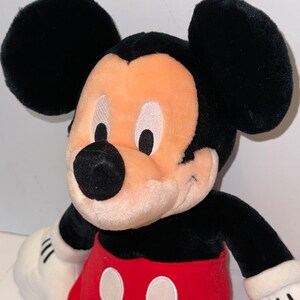 May include: A plush Mickey Mouse toy with black fur, large ears, and a red and white shorts. The toy has a smiling face with white eyes and a black nose.