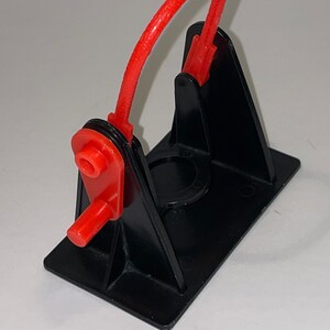 May include: A black plastic toy catapult with a red rubber band. The catapult has a base with a circular opening and a lever with a red handle.