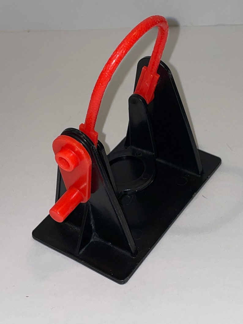 May include: A black plastic toy catapult with a red rubber band. The catapult has a base with a circular opening and a lever with a red handle.