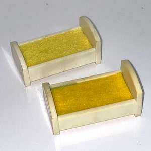 May include: Two white plastic doll beds with yellow fabric mattresses. The beds are small and designed for a dollhouse.