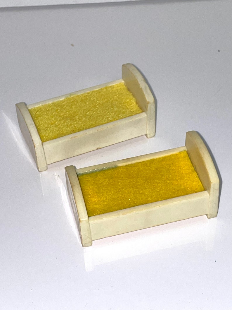 May include: Two white plastic doll beds with yellow fabric mattresses. The beds are small and designed for a dollhouse.