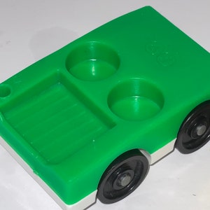 May include: Green plastic toy truck with a white stripe and black wheels. The truck has a bed in the back and two round holes in the top.