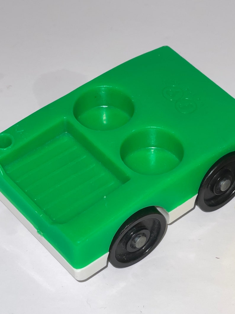 May include: Green plastic toy truck with a white stripe and black wheels. The truck has a bed in the back and two round holes in the top.