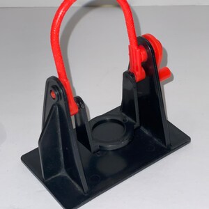 May include: A black plastic toy with a red plastic loop and a small red lever. The toy is designed to hold a small object in the centre.